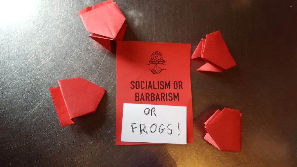 A table with five red cards. One reads SOCIALISM OR BARBARISM and the remaining text has been obscured by another piece of paper reading OR FROGS! This card is surrounded by four cards folded into paper frogs.